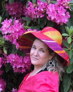 A picture of Kit dressed in bright pink and colorful hat in front of blooming rhododenron bush with bright pink flowers